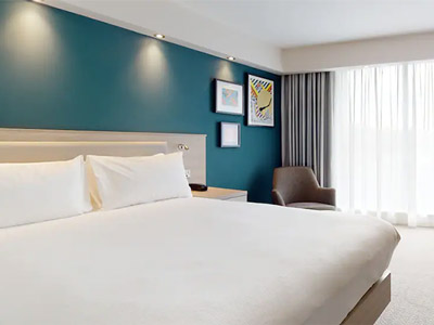 A double bed in a room with a blue wall and cushioned armchair in the corner at Hampton by Hilton Manchester Northern Quarter