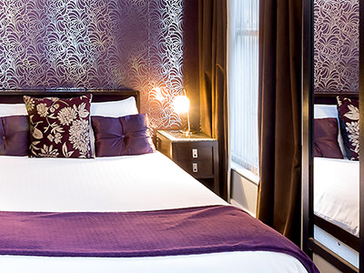 Image of a double bed with white linen and a purple accent throw, There is a bedside table with light and window. 