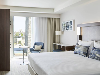Image of a large double bed with white linen and a window in the background with a table and chair in front of the window 