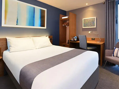 A spacious Travelodge super room with a double bed, desk space. a chair and lots of storage at Travelodge London Docklands