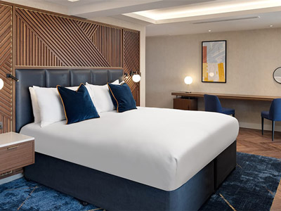 A blue themed double bed against a wooden wall with a long desk and two chairs at the side at Hilton London Metropole
