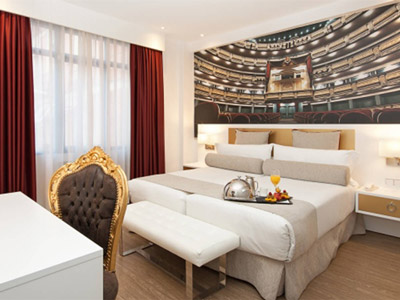 A king bed with an canvas of a theatre behind the bed and a desk and chair in front at Hotel Mayorazgo in Madrid 