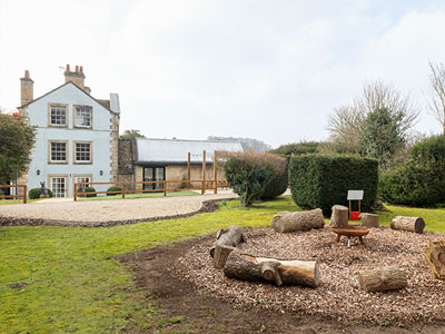 The outside of Cotswold Spa House with a spacious garden area including a circle of wooden seats made from logs 