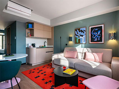 A living area that has a big pink sofa, pink wall art and a cabinet with a sink and tea/coffee making facilities 