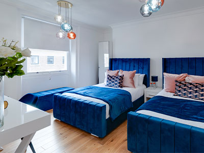 A bright bedroom at Rodney Street Apartments with two beds with blue headboards and covers 