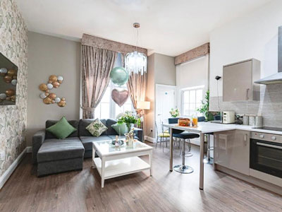 An open plan kitchen and lounge area that has an L shaped sofa, a wall mounted flatscreen TV and some decorative balloons 