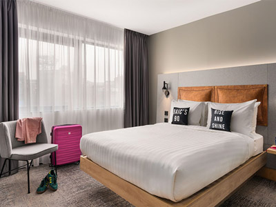 A Moxy hotel bedroom with a double bed and two slogan cushions one reading rise and shine and the other reading craic's 90 