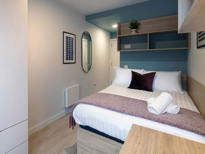 An apartment bedroom that has a double bed, shelf space above the bed and a mirror on the wall 