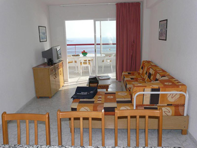A living room area with a sofa, tv and dining table facing out onto a sea view terrace that has a white table and chairs on it 