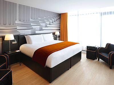 A double bed in front of a black and white mural on the wall, topped with a throw, with black chairs in the foreground