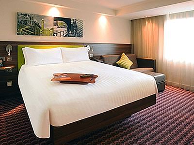 A white double bed in a hotel room, with a city skyline picture above the bed, with a brown leather sofa to one side of the bed