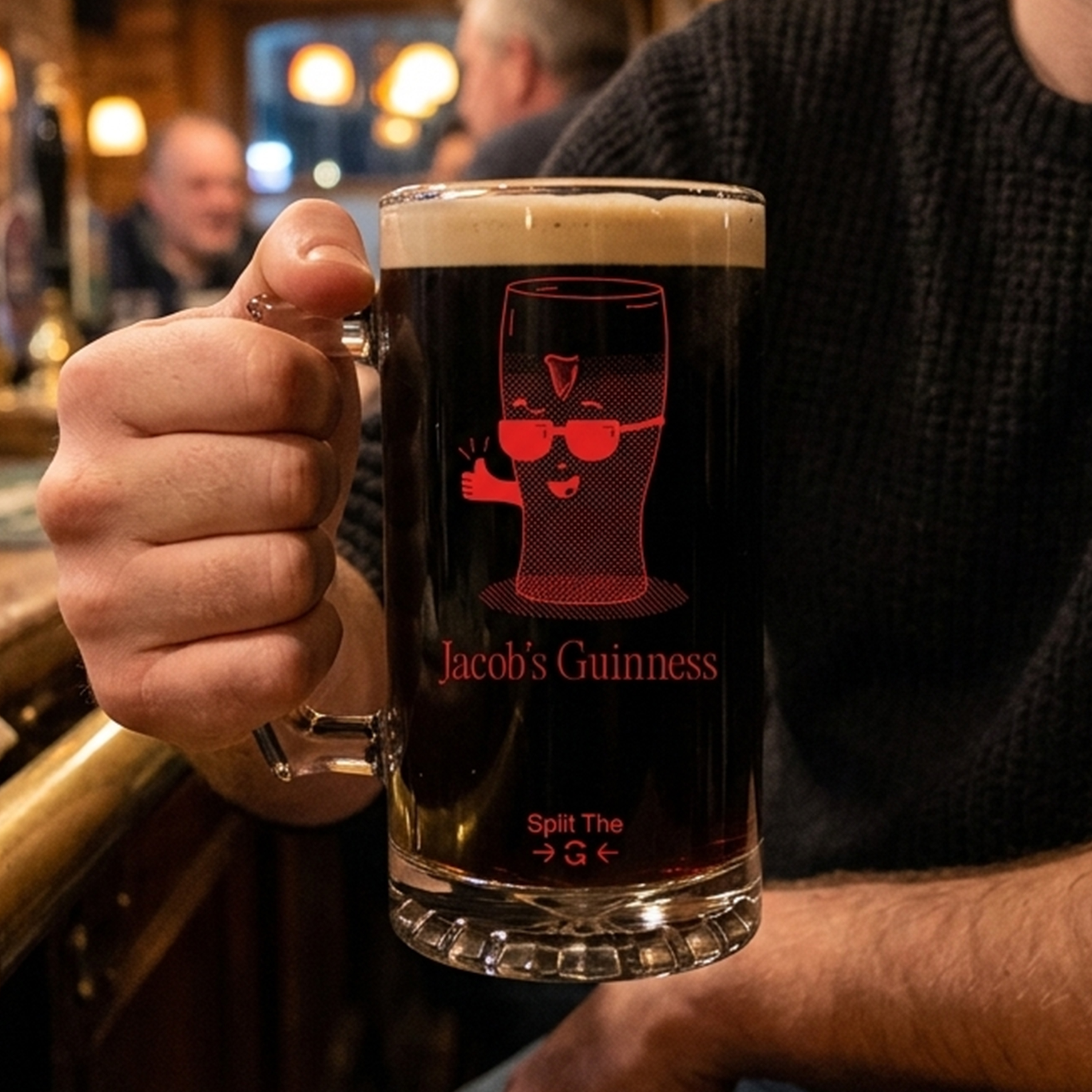 A man's hand holding a custom glass stein with a graphic of a pint of Guinness wearing glasses in red on it with the text Jacob's Guinness and towards the bottom of the stein it reads Split The G 