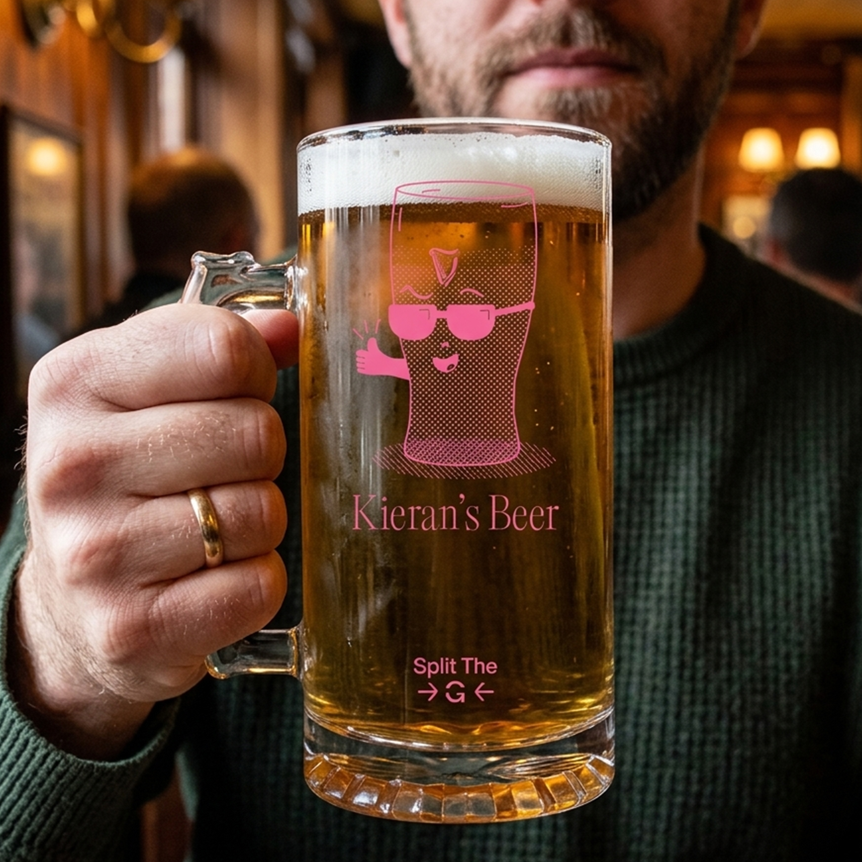 A man's hand holding a custom glass stein with a graphic of a pint of Guinness wearing glasses in red on it with the text Kieran's Beer and towards the bottom of the stein it reads Split The G