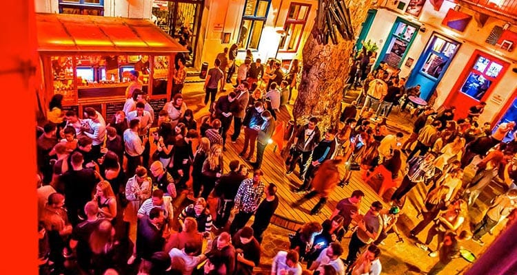 Image of people standing in a massive open bar with a big tree in the middle of the room and multi coloured doors and windows