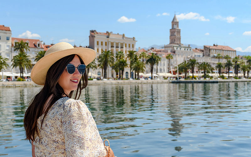 A brunette woman in a sun hat and glasses posing by Splits seafront. 