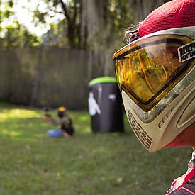 A man in the foreground wearing a protective visor, with an outdoor paintball arena in the background