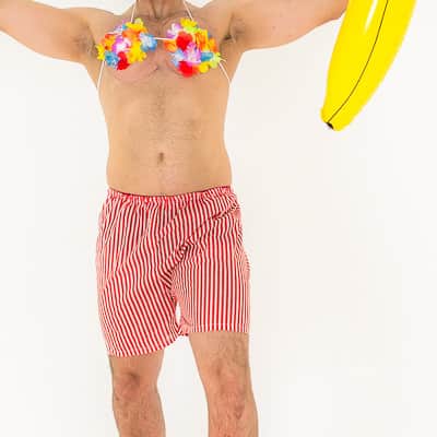 A man in a Hawaiian Lei bra and beach shorts, holding up an inflatable banana and champagne bottle with a beachball in front
