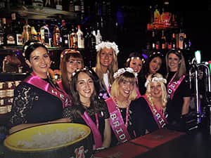 Women on a hen weekend, stood behind a bar and wearing hen party sashes