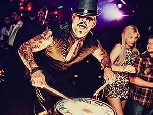 A man in a top hat and waistcoat playing the drums in a busy club 