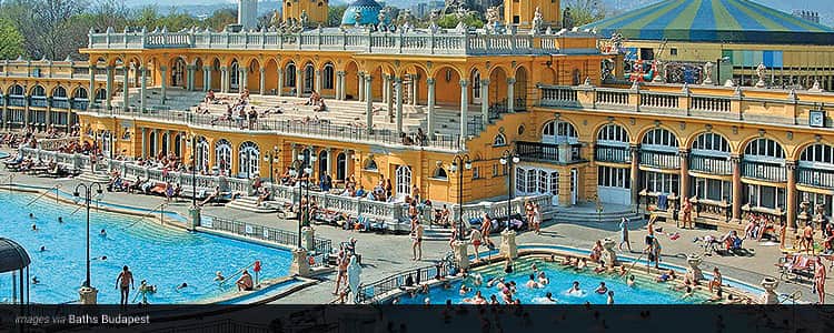 The yellow exterior and outdoor pool of the Turkish Baths, Budapest, with people standing outside of the building and in the pool