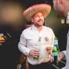 A man wearing a sombrero and holding a bottle and glass of white wine, chatting to another man