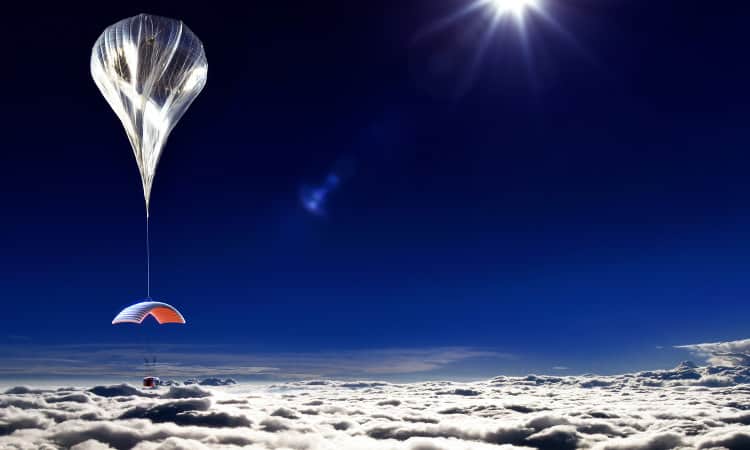 Balloon rising into the blue sky with clouds below and the sun rising high in the sky.