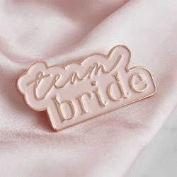 A pastel pink enamel badge with Team Bride written in rose gold on a pale pink piece of fabric.