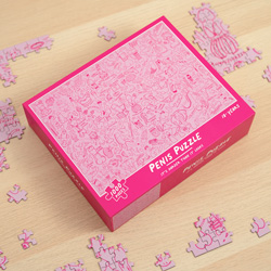 Penis Jigsaw Puzzle box with pieces spread around on a wooden table.