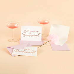 Will You Be My Bridesmaid card and envelopes and some cocktails on a yellow background.